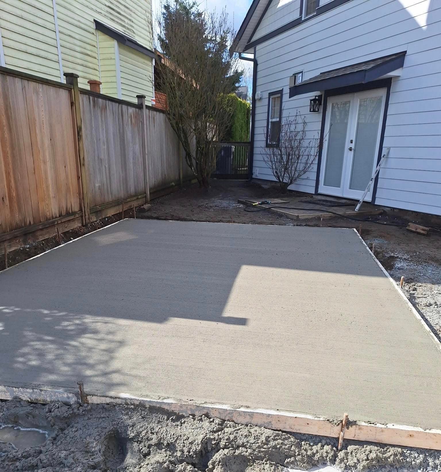 Freshly poured concrete slab in a backyard, ready for construction with surrounding wooden fence.