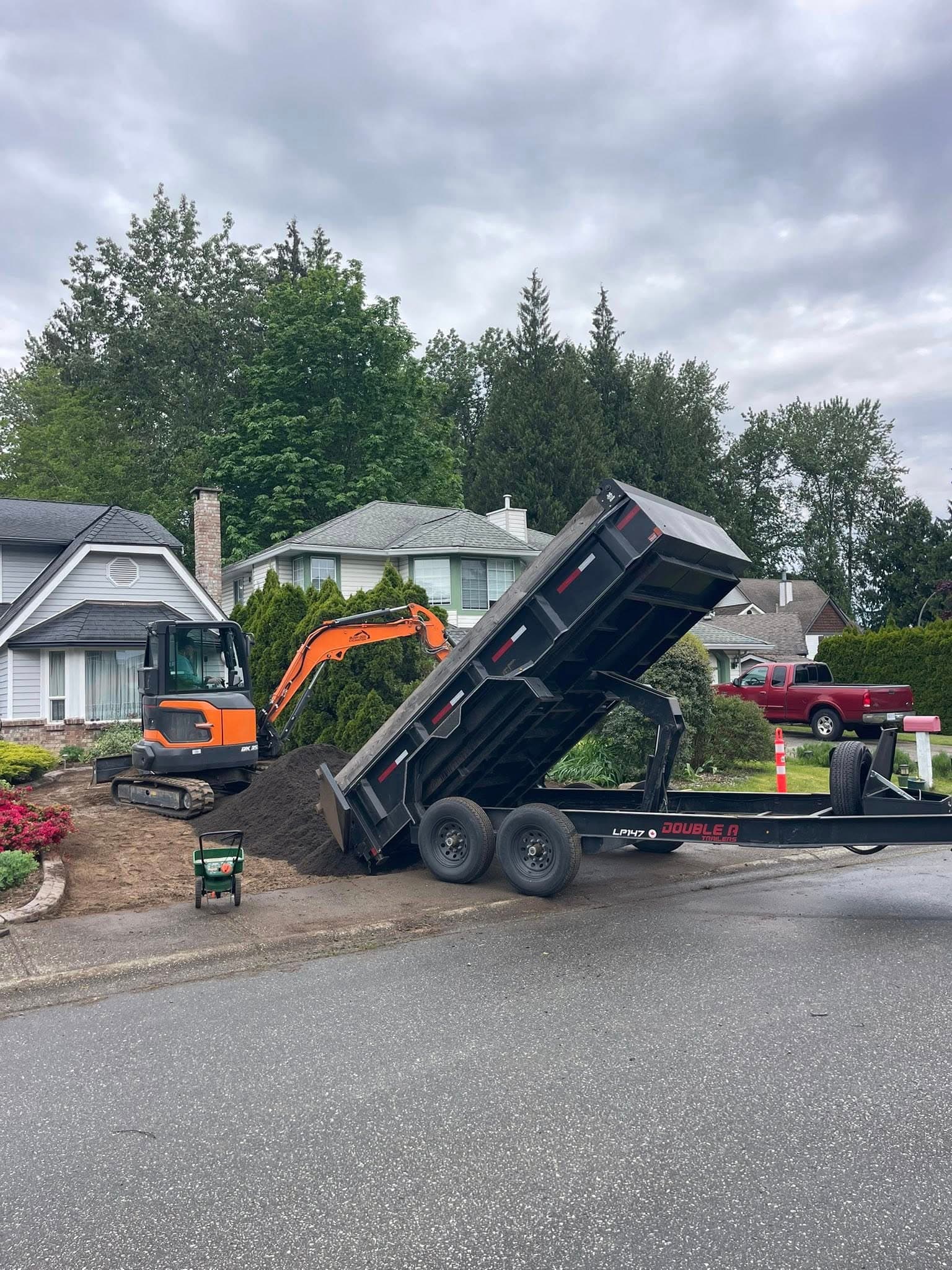 Dump truck unloading dirt near a house with construction equipment and greenery.